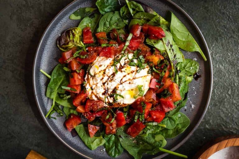 A plate of fresh spinach, diced tomatoes, and a ball of burrata cheese drizzled with balsamic glaze.