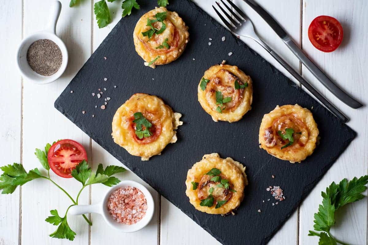 A slate platter with five mini pizzas garnished with parsley next to utensils. Surrounding the platter are bowls of pink salt and pepper, sliced tomatoes, and parsley sprigs on a white wooden table.