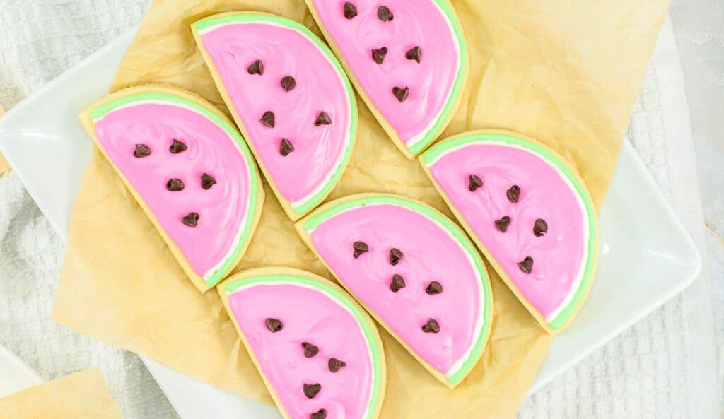 Cookies decorated to resemble watermelon slices with pink frosting, green borders, and chocolate chips as seeds, arranged on parchment paper.