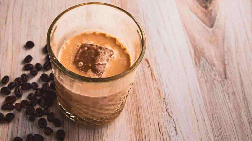 A glass filled with iced coffee sits on a light wooden surface, surrounded by scattered coffee beans.