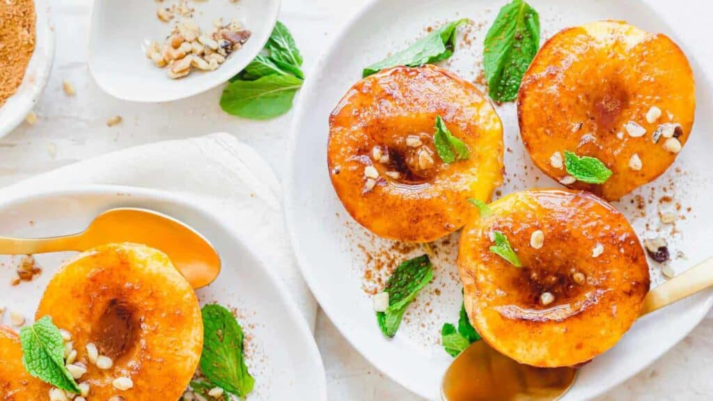 Three caramelized peach halves garnished with fresh mint and nuts on a white plate with a spoon on the side.