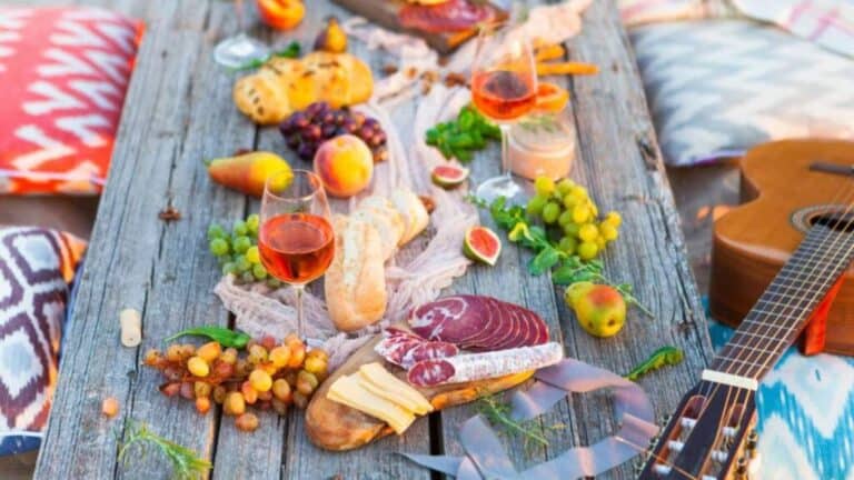 A rustic wooden table is set with a variety of foods, including bread, fruits, cheese, and charcuterie, accompanied by two glasses of rose wine. Colorful pillows and a guitar lie in the background.