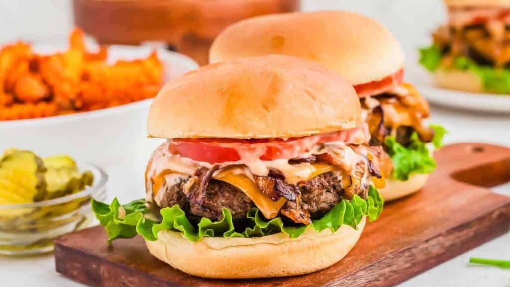 Two cheeseburgers with lettuce, tomato, caramelized onions, and sauce on buns are served on a wooden board. A bowl of pickles and a plate of sweet potato fries are in the background.