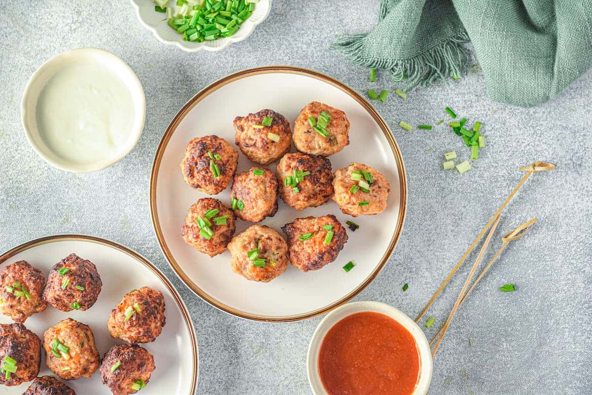 Plates of meatballs garnished with chopped green onions are placed next to dipping sauces and a gray napkin on a light-colored surface.