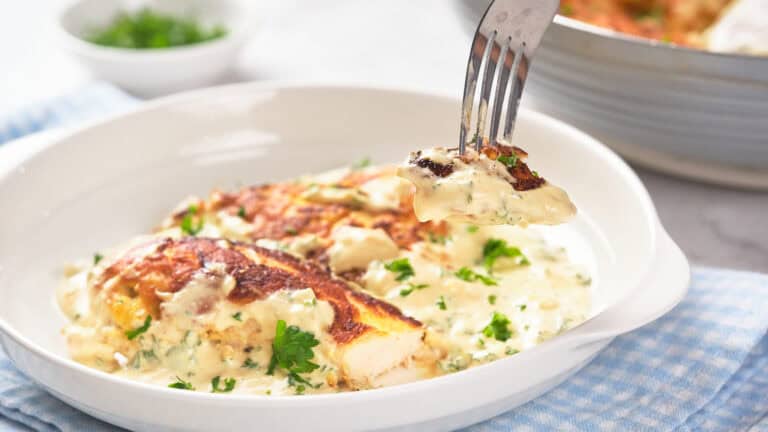 A fork holds a piece of creamy, herbed chicken above a white plate containing more pieces in a white sauce. Chopped parsley garnishes the dish. A small bowl with garnish is blurred in the background.