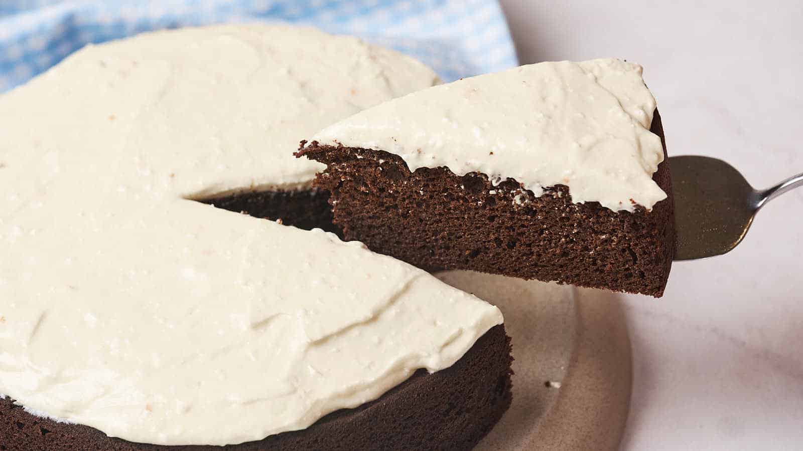 A slice of chocolate cake with white frosting being lifted from the whole cake.