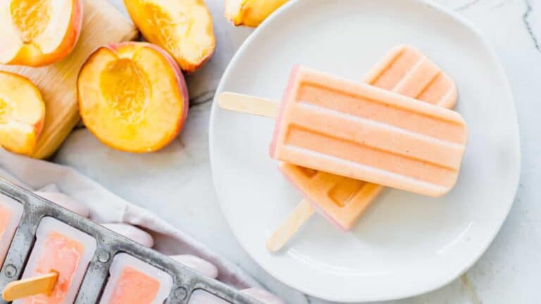 Two peach popsicles on a white plate, surrounded by sliced peaches and a popsicle mold.