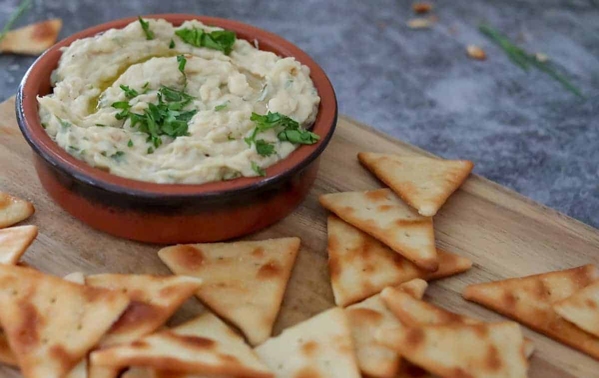 A small bowl of creamy dip garnished with herbs is placed on a wooden board surrounded by triangular-shaped pita chips.