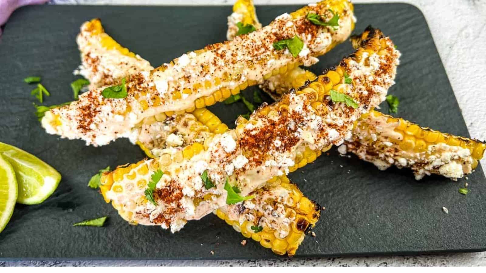 Grilled corn on the cob pieces topped with cheese, chili powder, and cilantro, served on a black slate plate with lime wedges on the side.