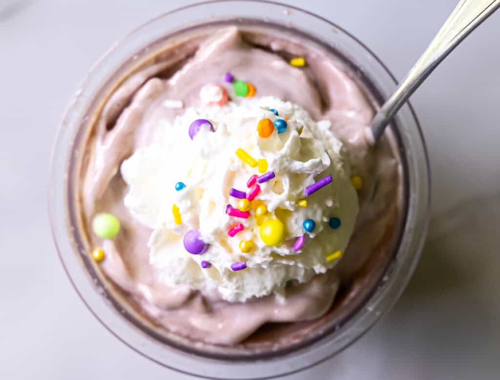 A dessert cup filled with chocolate mousse topped with whipped cream and colorful sprinkles, with a spoon in it.