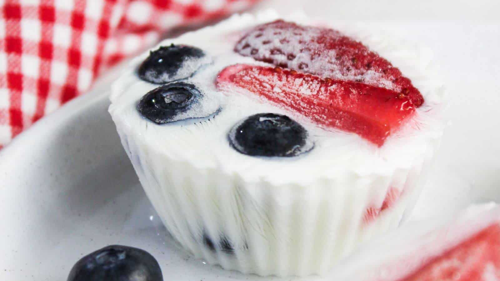 A frozen yogurt cup with blueberries and a strawberry slice on top, placed on a white plate. A red and white checkered cloth is visible in the background.