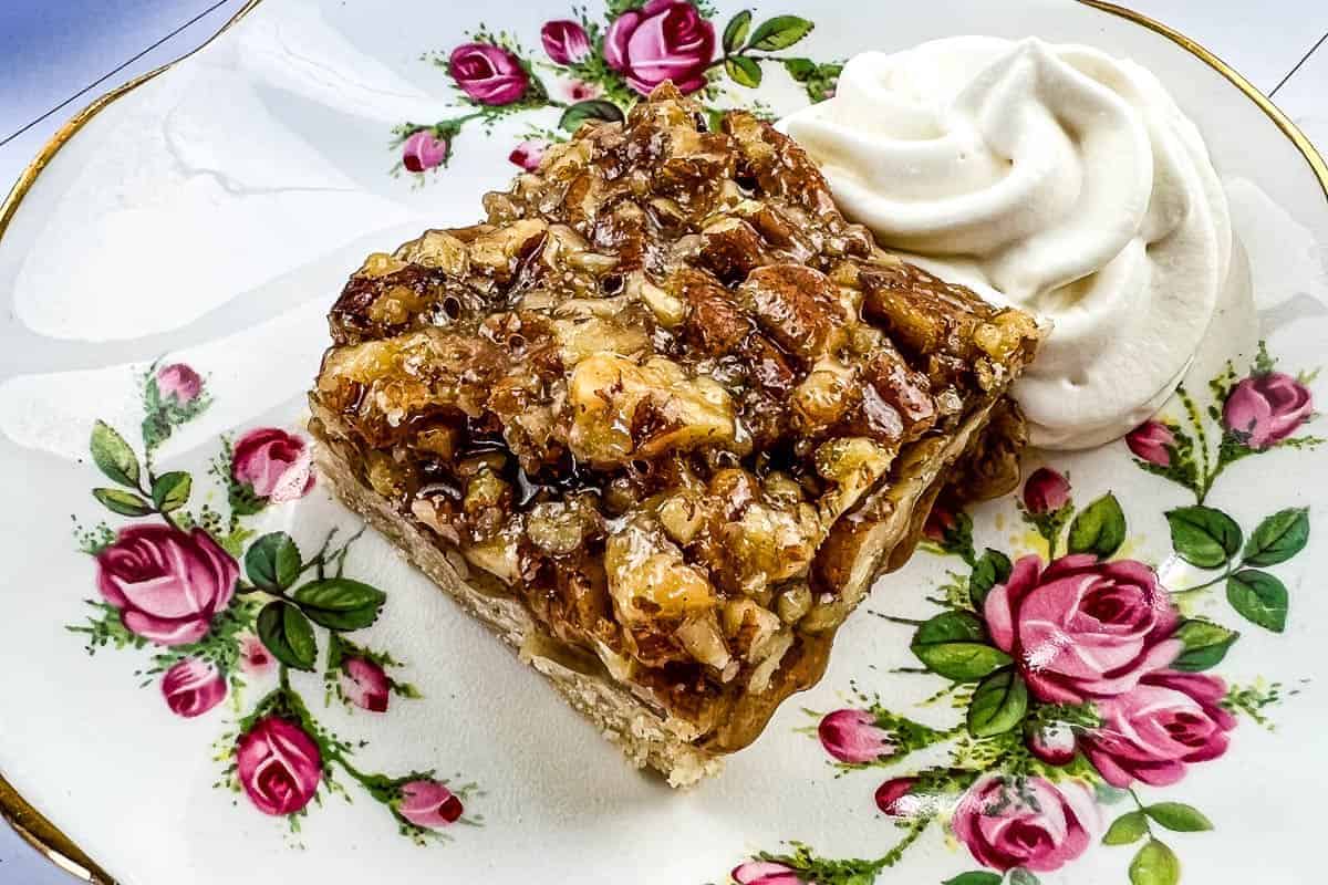 A slice of maple pecan pie bar topped with chopped pecans sits on a floral-patterned plate.