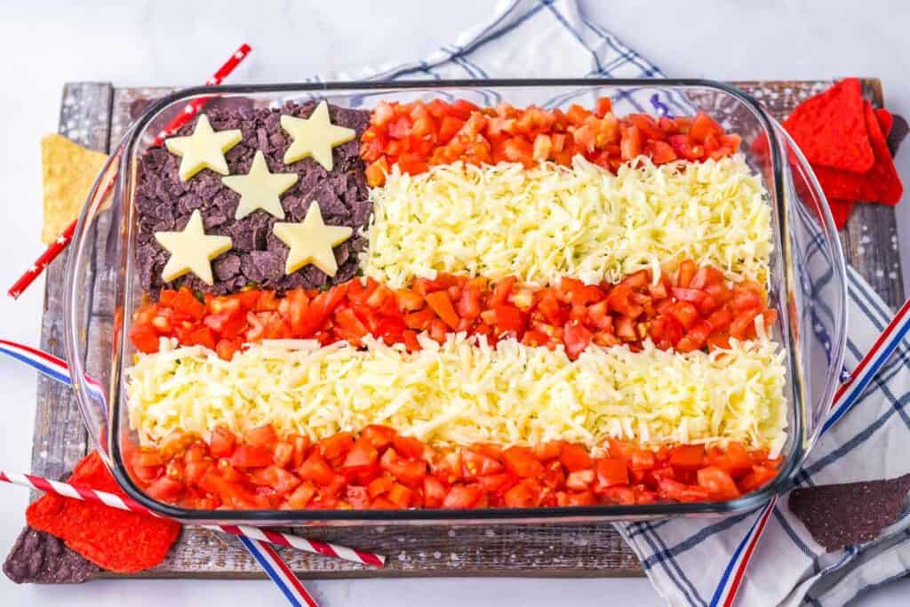 A rectangular dish of layered dip arranged to resemble the American flag, with cheese and tomatoes for stripes, and stars on blue corn chips for the stars.