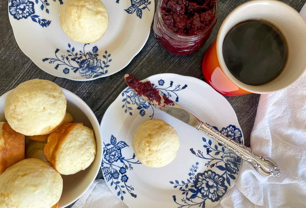 A table with plates of scones, a jar of jam, a butter knife with jam spread, and a cup of coffee.