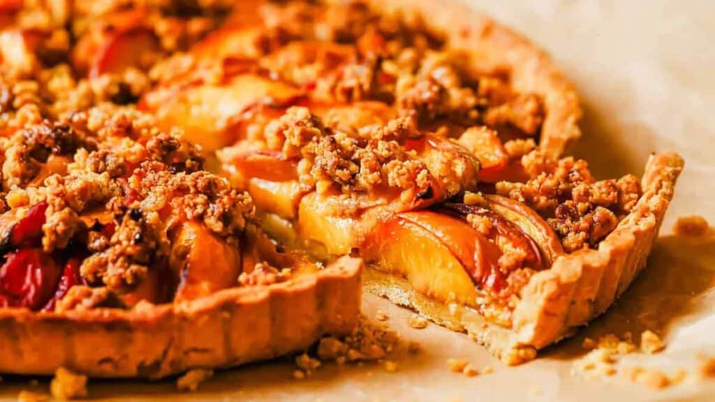 Close-up of a peach crumble tart on parchment paper, with a slice partially removed, revealing a flaky crust and a filling of peach slices topped with crumbly streusel.