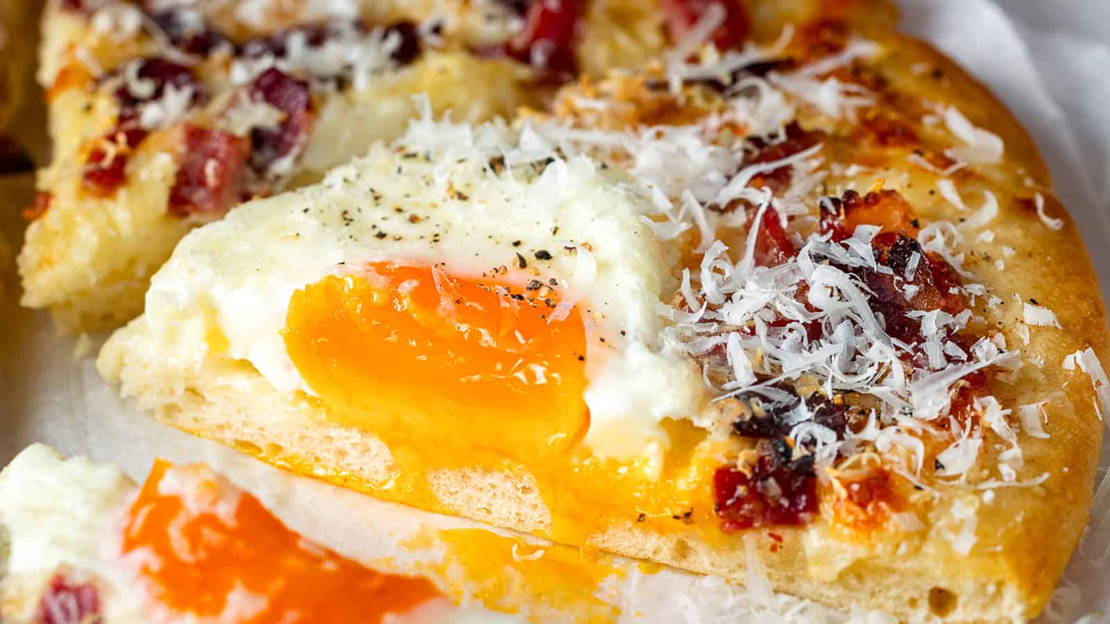 A close-up of a pizza slice topped with a runny egg yolk, grated cheese, bacon bits, and black pepper.
