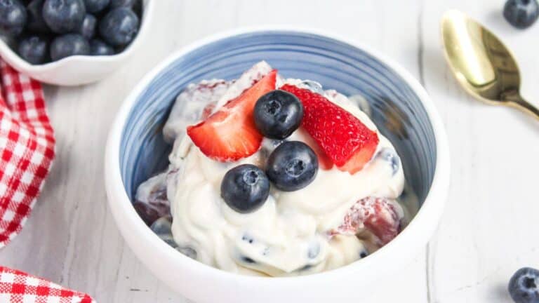 A bowl of creamy berry salad topped with fresh blueberries and sliced strawberries. A gold spoon is placed beside the bowl, and there is a small dish of blueberries in the background.