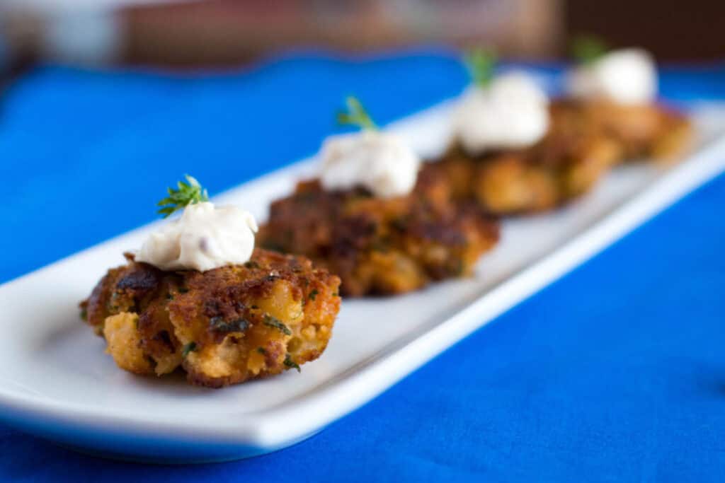 A plate with several small, browned crab cakes, each topped with a dollop of white sauce and a garnish of fresh herbs, placed on a blue tablecloth.