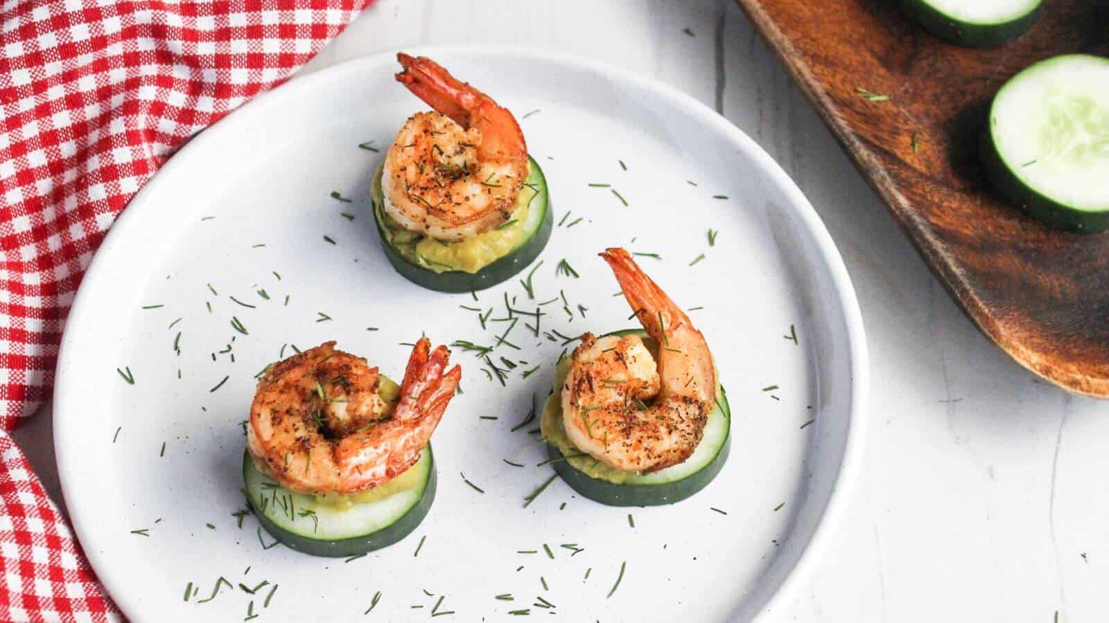 Three grilled shrimp atop cucumber slices garnished with herbs on a white plate with a red checkered napkin partially in view.