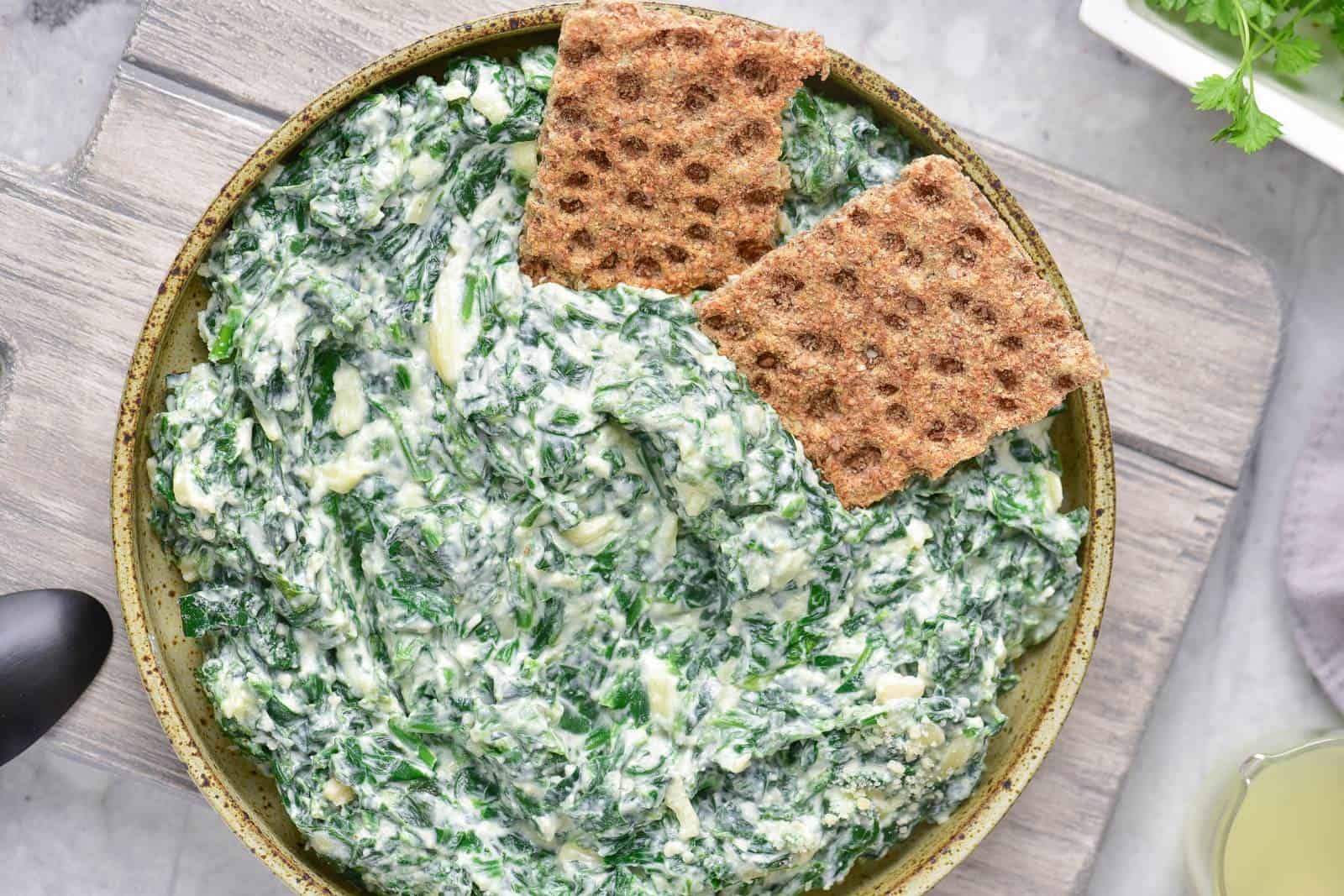 A bowl of creamy spinach artichoke dip with two pieces of crispbread on top. The bowl is placed on a wooden board.