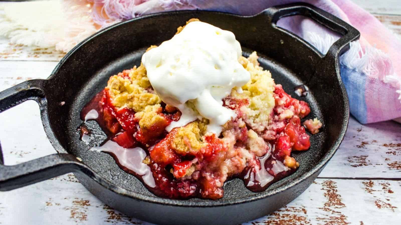 A cast iron skillet with strawberry rhubarb cobbler topped with a scoop of vanilla ice cream on a wooden surface with a cloth napkin beside it.