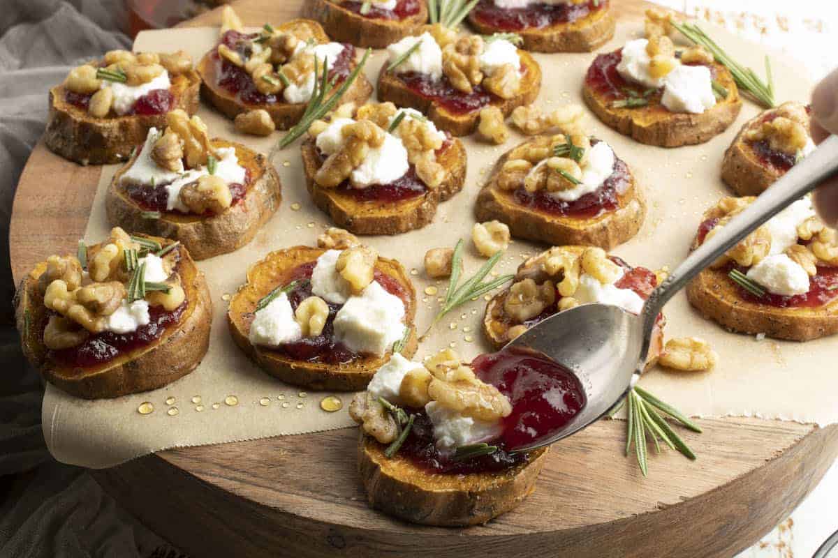 A wooden tray holds numerous sweet potato rounds topped with cheese, cranberry sauce, walnuts, and fresh rosemary, with a spoon drizzling honey over one of the rounds.