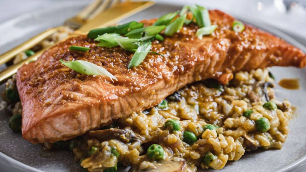 A plated dish featuring a seared salmon fillet topped with chopped green onions, served over a bed of risotto with peas and mushrooms. A fork and knife are placed beside the salmon.