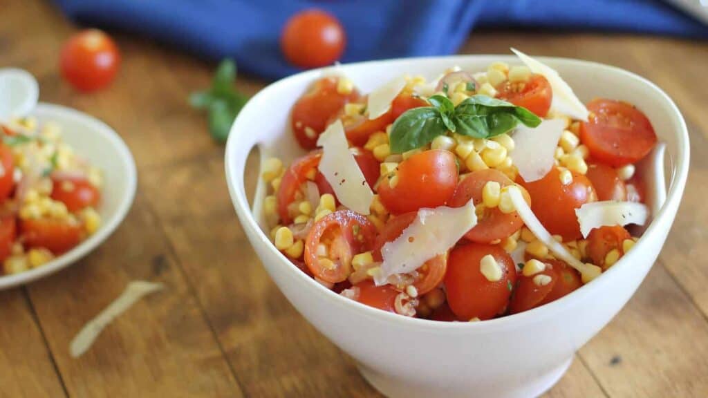 A white bowl filled with a salad consisting of cherry tomatoes, corn, shaved cheese, and garnished with a basil leaf. Another bowl with similar salad and a few cherry tomatoes are in the background.