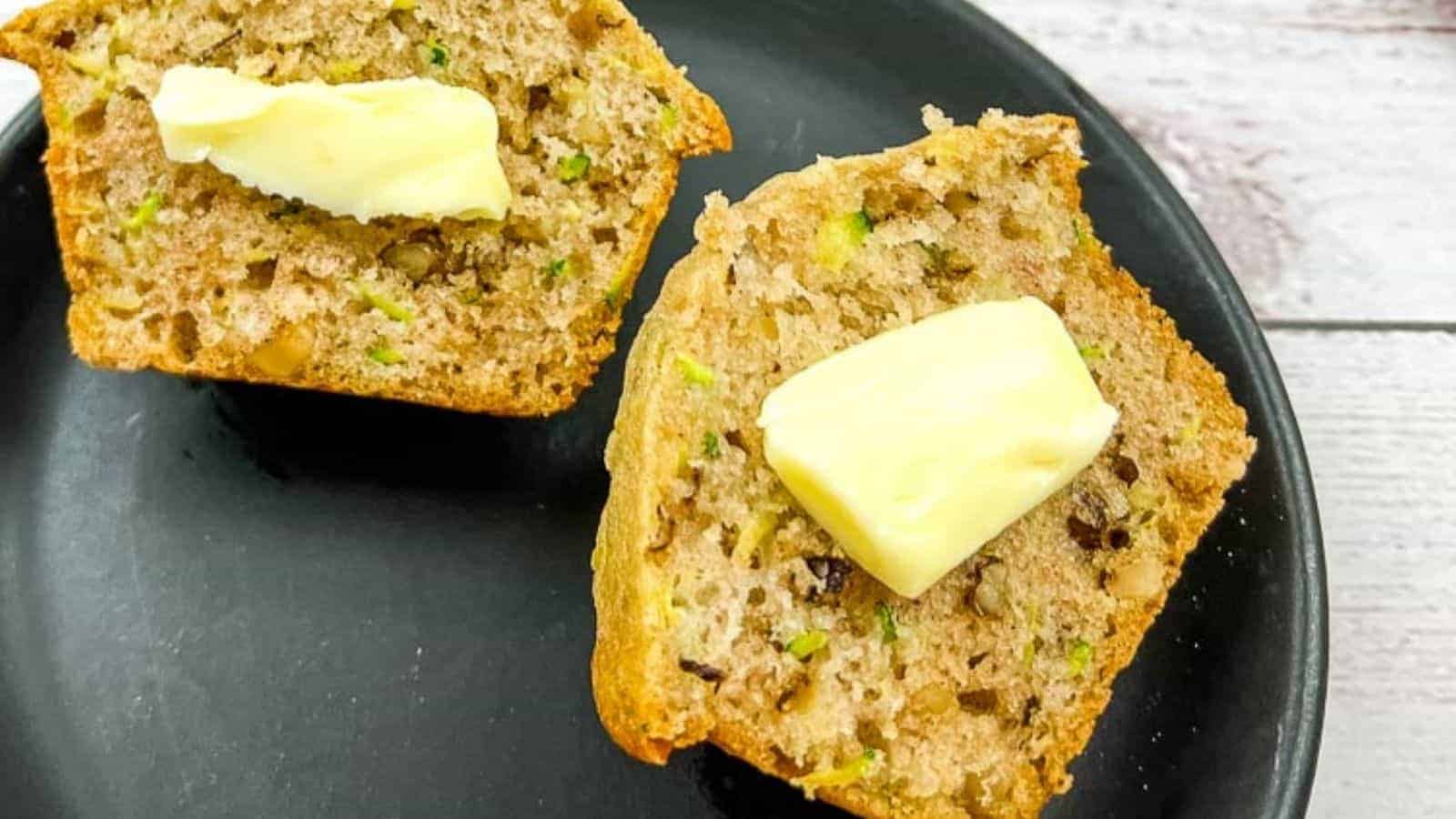 A sliced muffin with visible nuts and green specks is topped with butter and placed on a black plate.