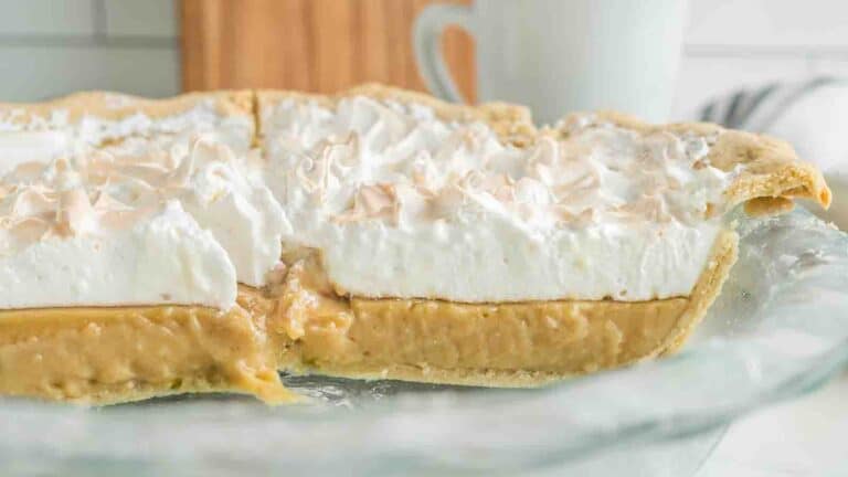 A partially sliced pie with a golden crust, filled with a layer of yellow custard and topped with fluffy white meringue on a glass plate.