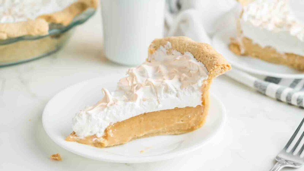 A slice of cream pie with a golden crust and whipped topping sits on a white plate, next to a full pie and a white cup in the background.