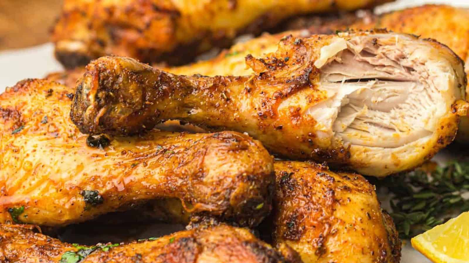 Close-up of a stack of seasoned, grilled chicken drumsticks, with one drumstick partially eaten, revealing moist, cooked meat inside. A lemon wedge and herbs are visible in the background.