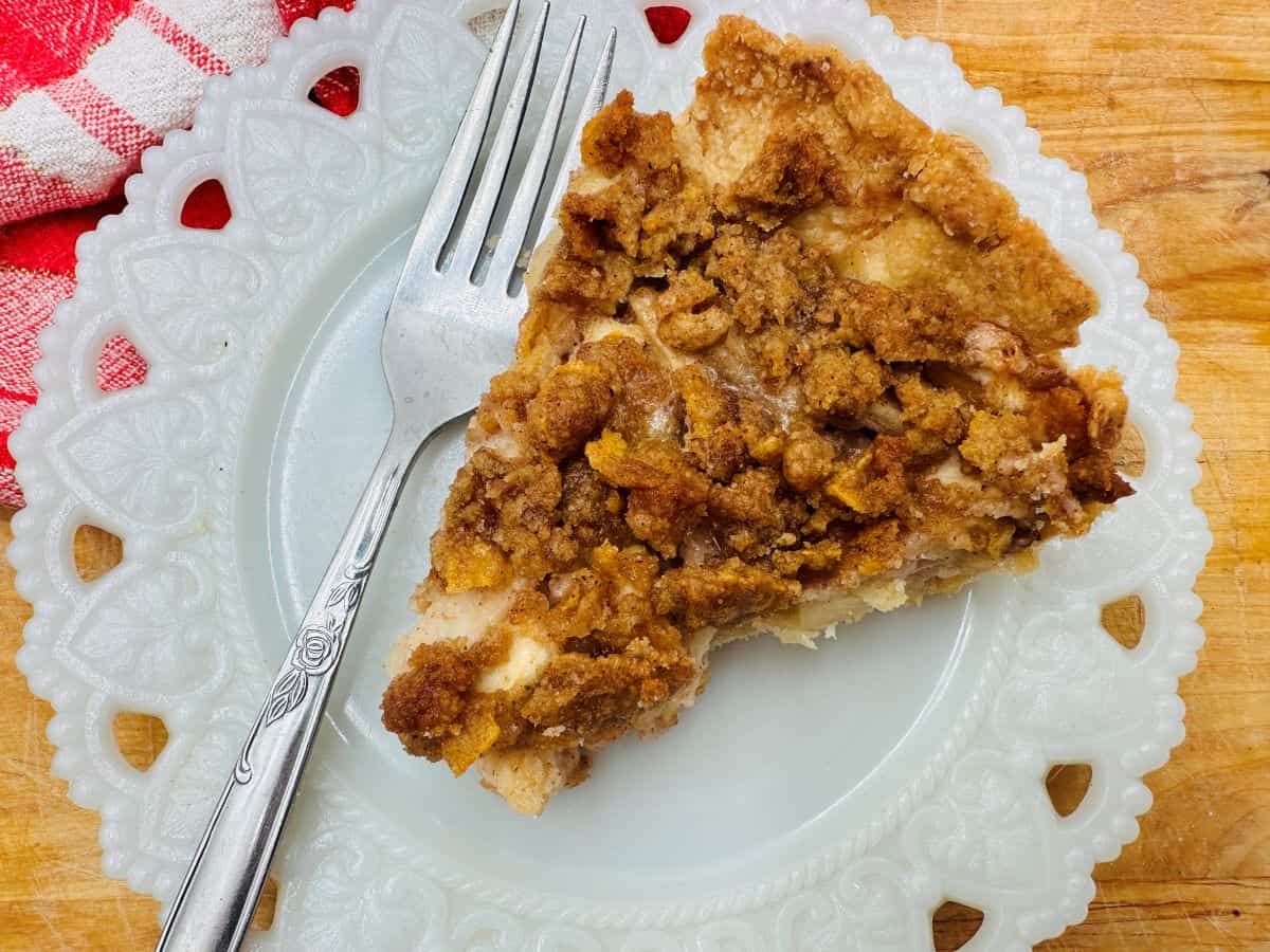 A slice of crumb-topped pie on a white decorative plate with a fork next to it. A red and white cloth is partially visible in the corner.