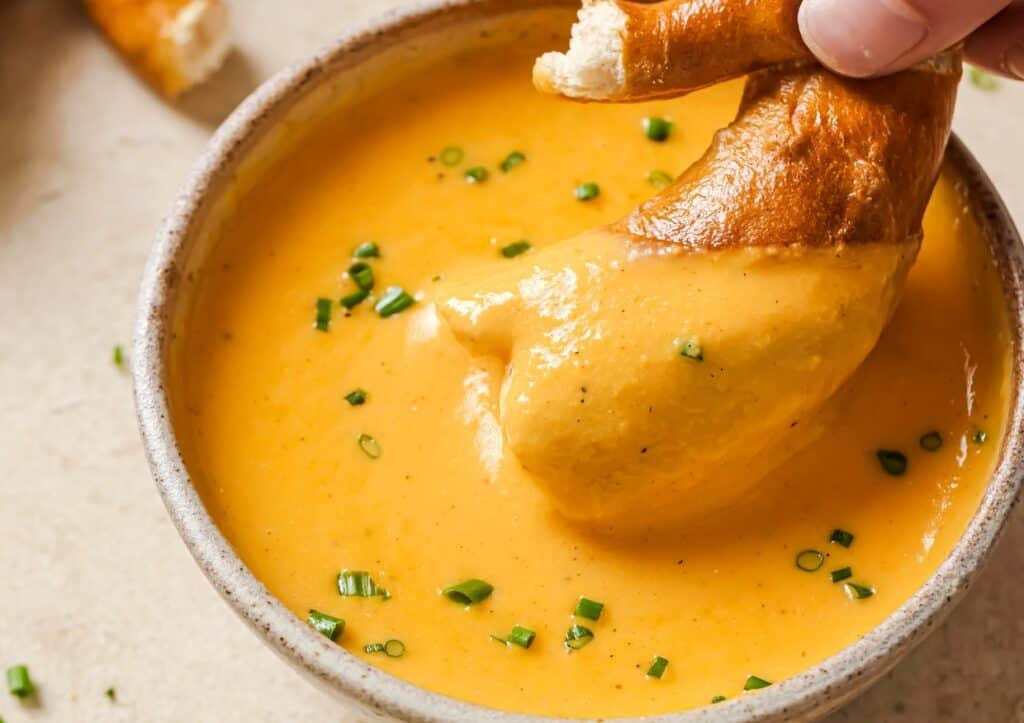 A hand dips a pretzel into a bowl of creamy cheese sauce garnished with chopped chives.