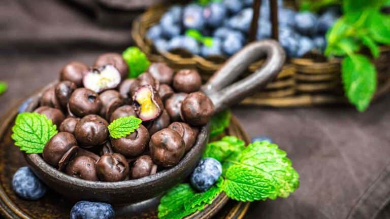 Chocolate-covered blueberries in a rustic bowl, garnished with mint leaves, with a similar basket of blueberries and mint in the background.