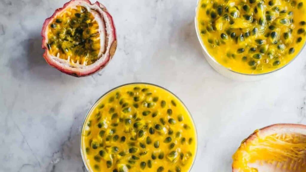 Two glasses filled with yellow passion fruit mousse, placed beside cut open passion fruits on a light-colored surface.