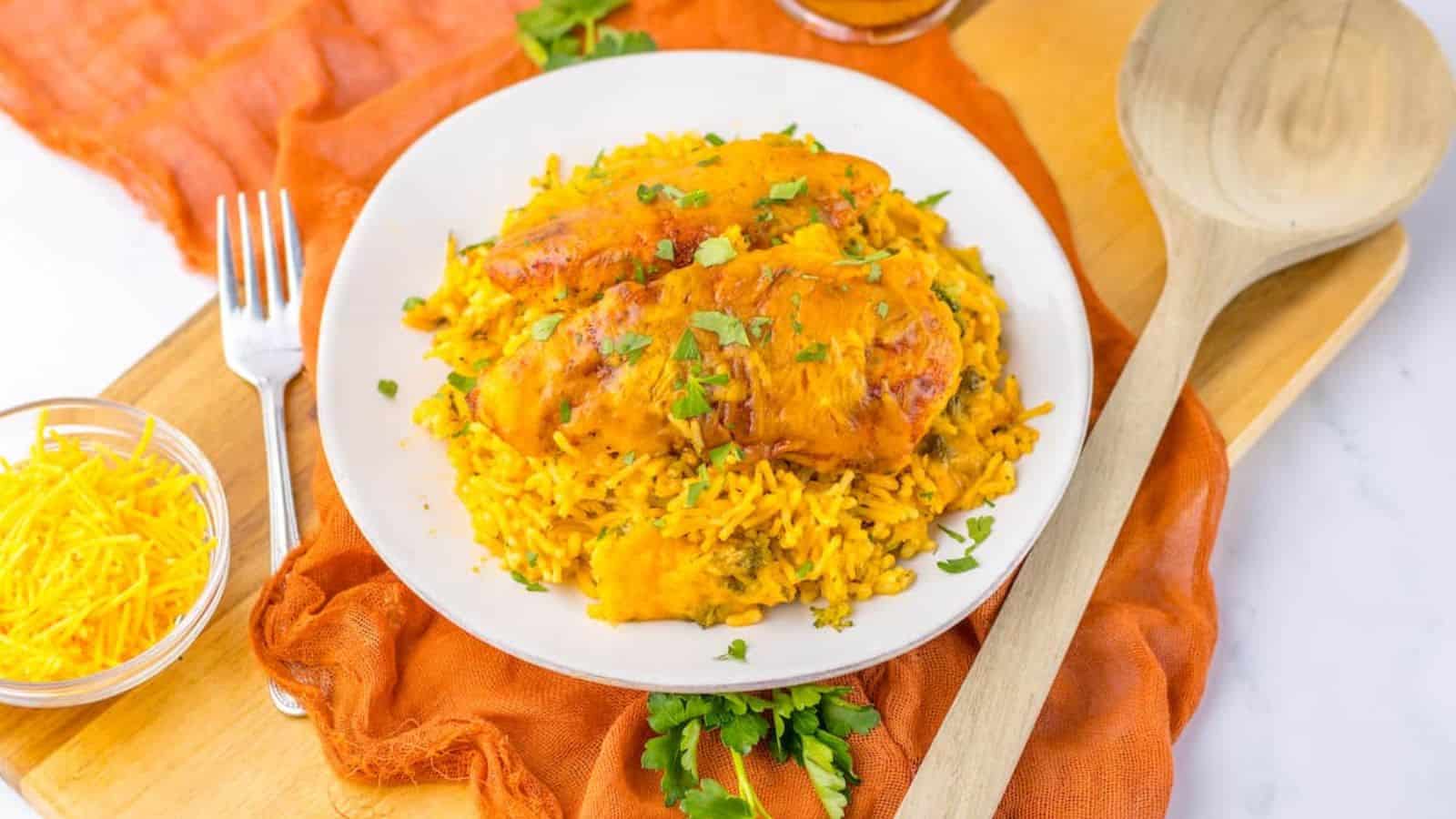 A plate of chicken with rice garnished with parsley on a white surface with an orange napkin, wooden utensils, and ingredients nearby.