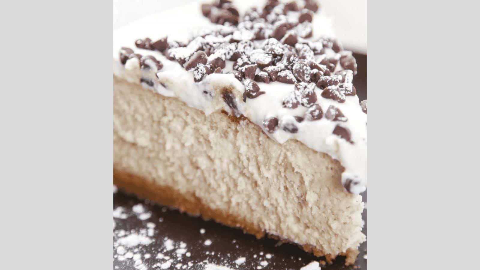 A slice of cannoli cheesecake topped with mini chocolate chips and dusted with powdered sugar.