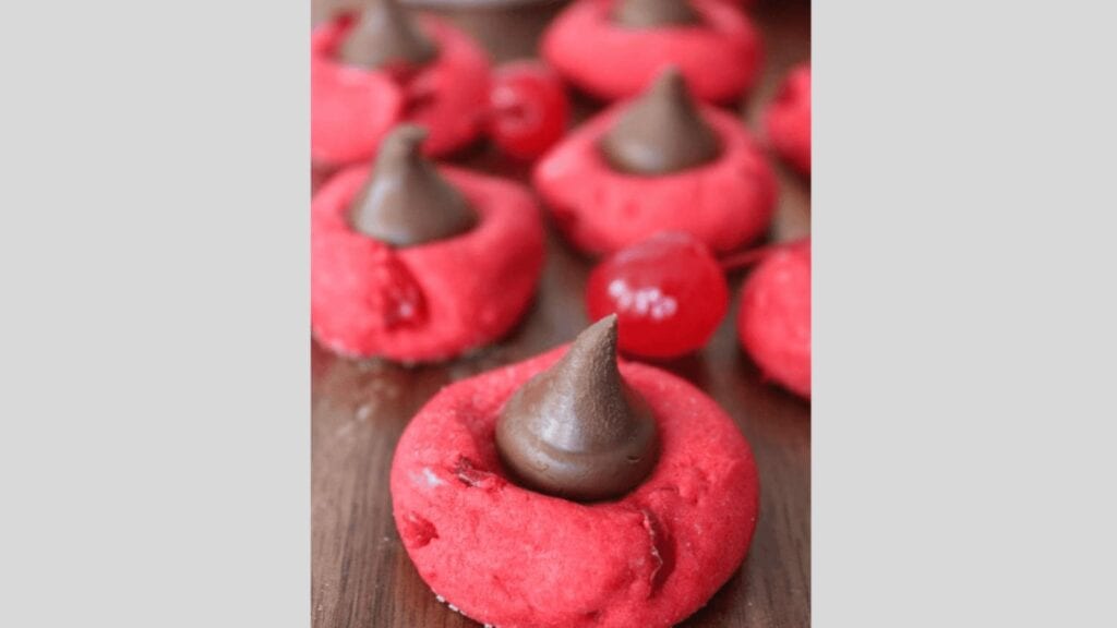 Close-up of red thumbprint cookies topped with chocolate kisses on a wooden surface, with maraschino cherries arranged in the background.