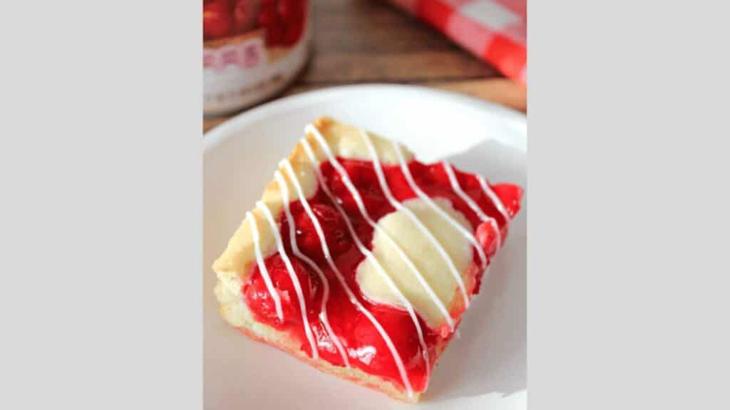 A piece of cherry Danish pastry with white icing drizzle, served on a white plate.