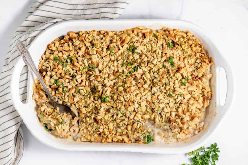 A white baking dish filled with a casserole topped with a golden-brown breadcrumb crust. A serving spoon rests inside the dish. A striped cloth napkin is placed beside it.