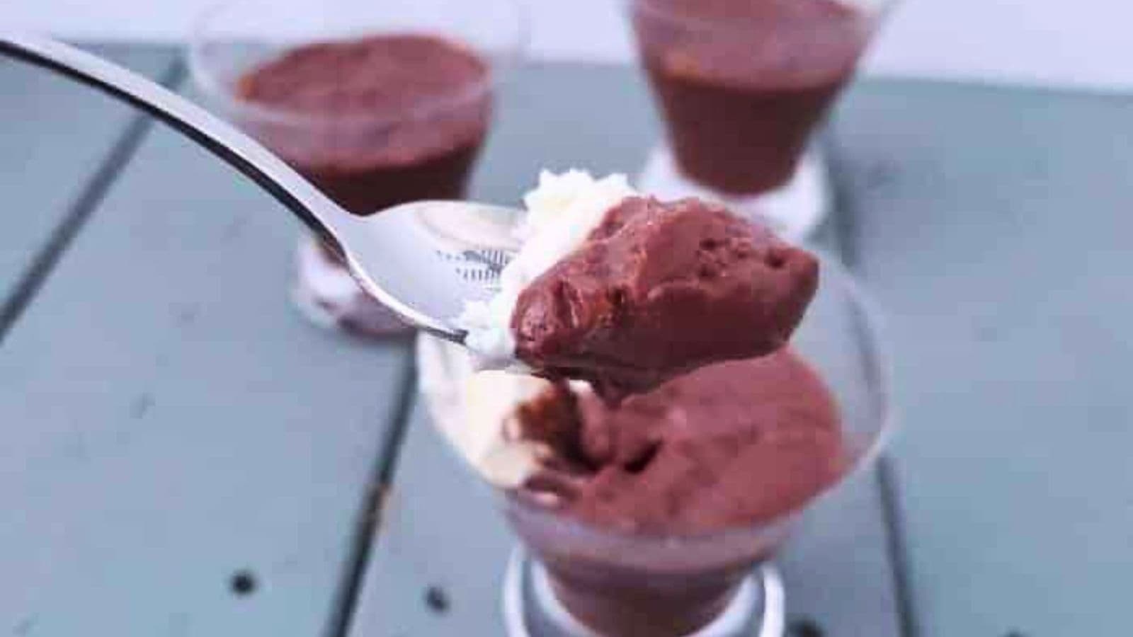 Image shows A close-up of a spoon with chocolate pudding above a glass filled with the same pudding. Three more glasses of pudding are in the background on a gray surface.