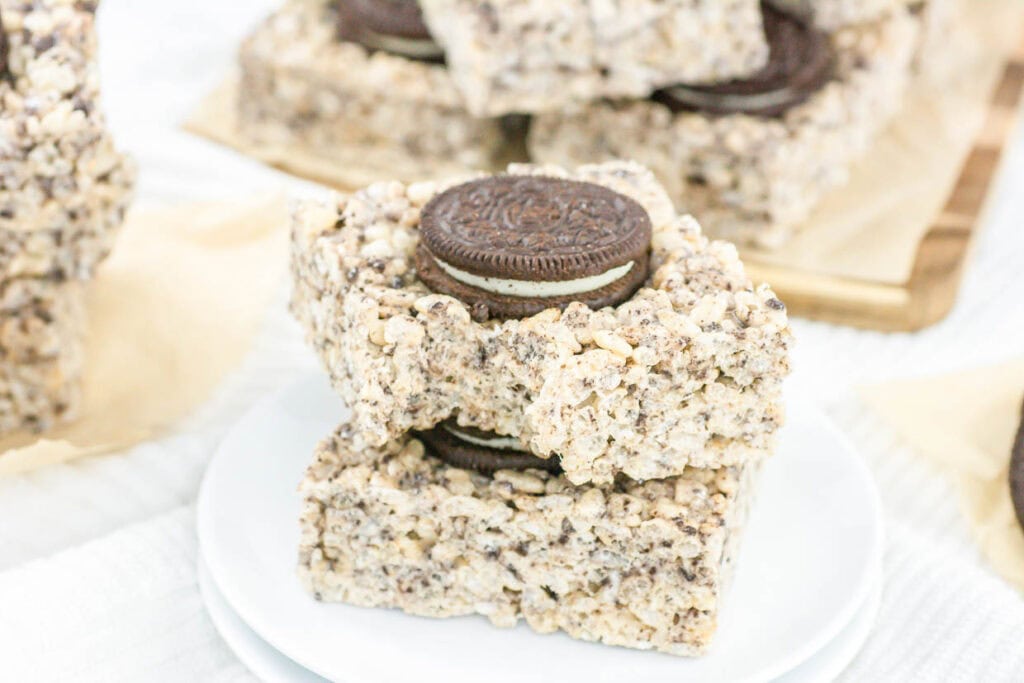 Two square rice cereal treats with a chocolate sandwich cookie in the center are stacked on a white plate. More of these Chill Out Desserts are visible in the background.