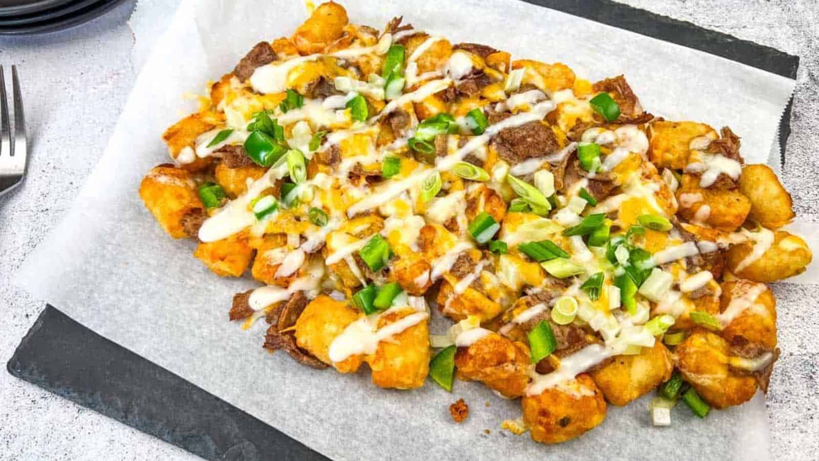 Philly Cheesesteak Loaded Tots on parchment paper, topped with Alfredo cheese, green onions, and a drizzle of white sauce, placed on a dark rectangular slate plate.