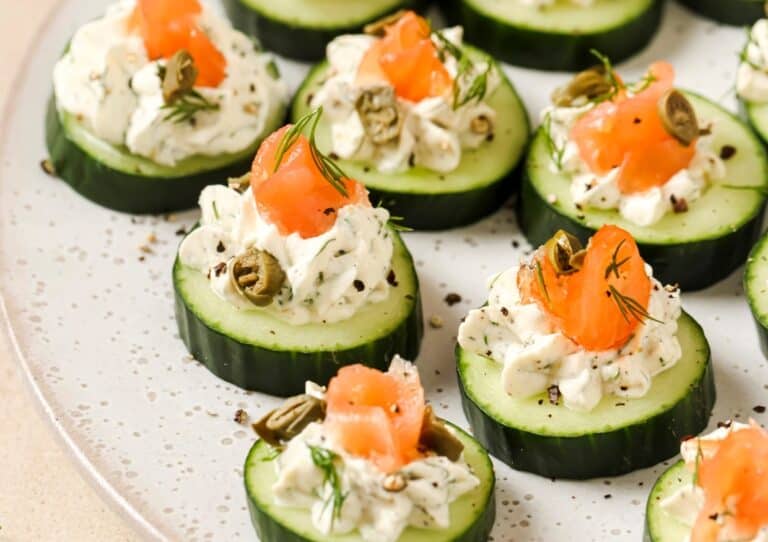 Cucumber slices topped with cream cheese, smoked salmon, capers, and dill are arranged on a plate.