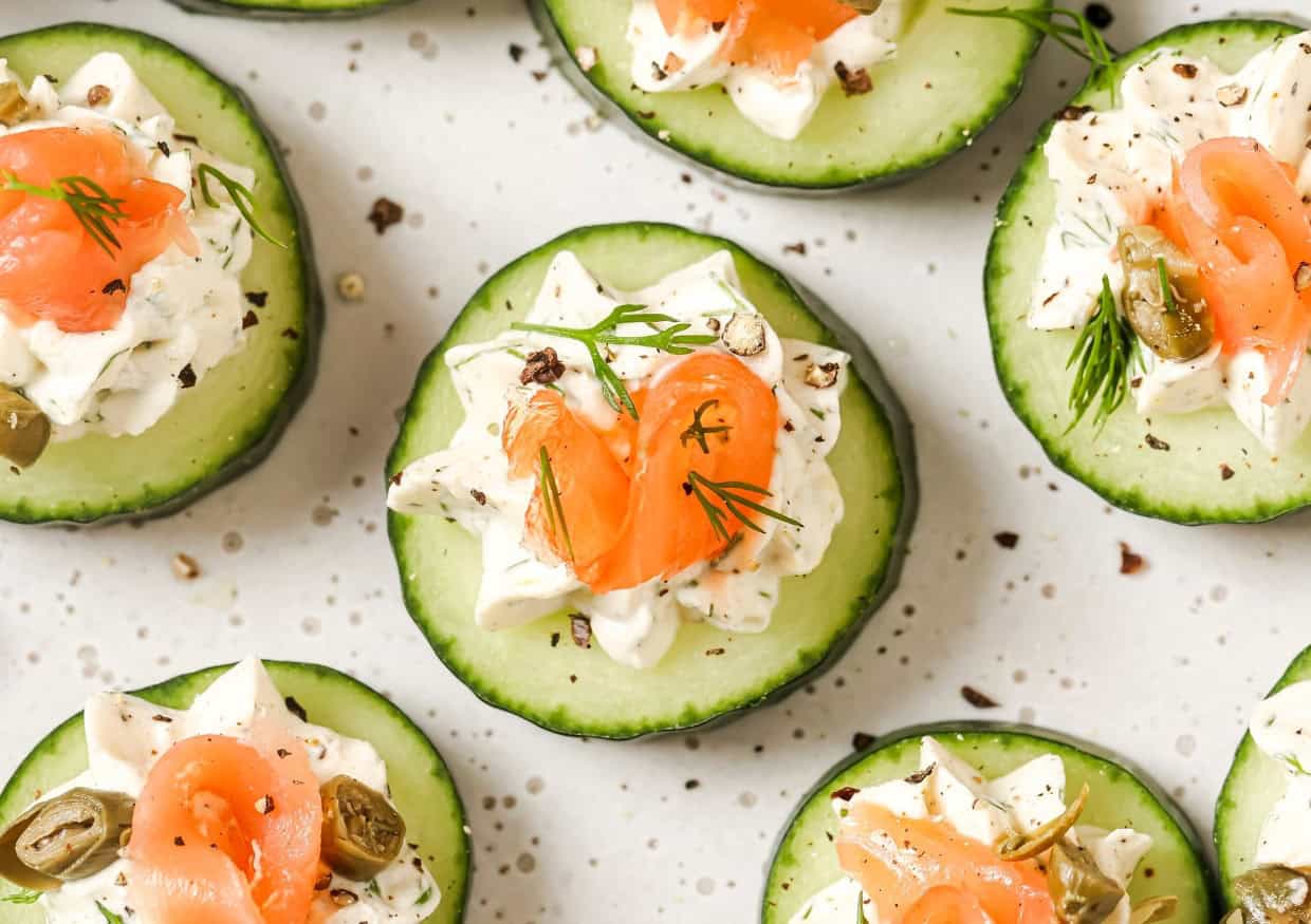 Cucumber slices topped with cream cheese, smoked salmon, capers, and dill, arranged on a white speckled surface.