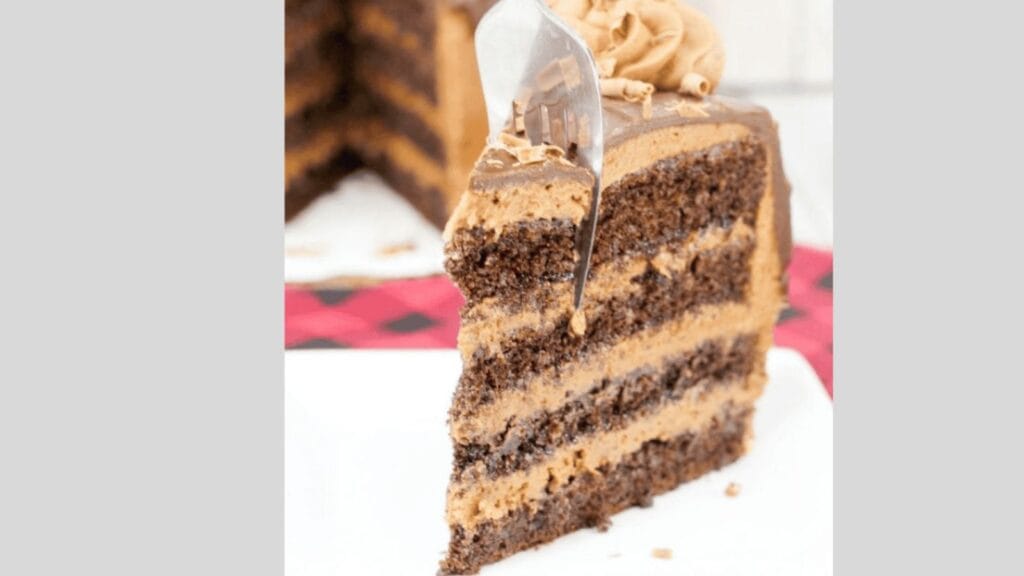 A close-up of a slice of chocolate layer cake with light brown frosting and chocolate shavings on top. A fork is inserted into the slice.