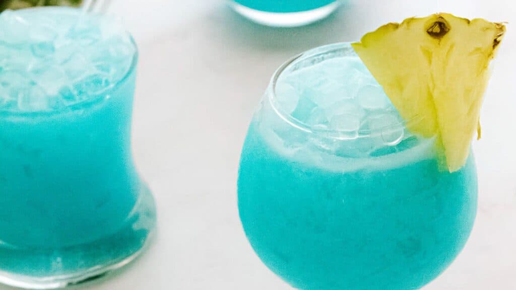 A close-up of a turquoise cocktail in a round glass, garnished with a pineapple slice. Two similar summer beverages are visible in the background.