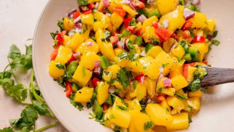 A bowl of colorful mango salsa with diced mango, red onions, red and green bell peppers, cilantro, and seasonings, stirred with a wooden spoon. Fresh cilantro garnish on the side.