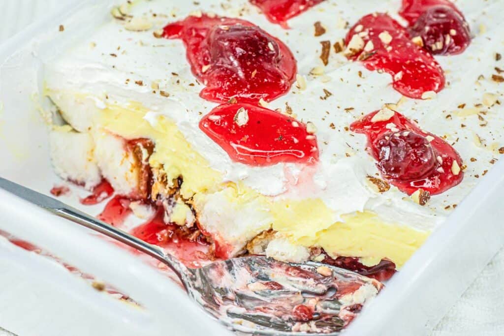 A white dish with a layered dessert consisting of whipped cream, strawberry topping, chopped nuts, and a yellow layer beneath. A metal spoon is placed in the dish with some dessert scooped out.